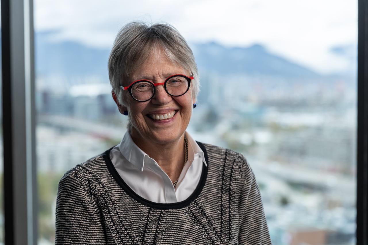 Portrait of Penny Ballem next to a window. She is wearing a grey sweater and white collared shirt