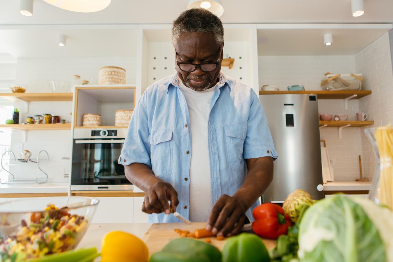 Senior person standing in the kitchen and chopping carrot on wooden board. He is preparing a healthy dinner.