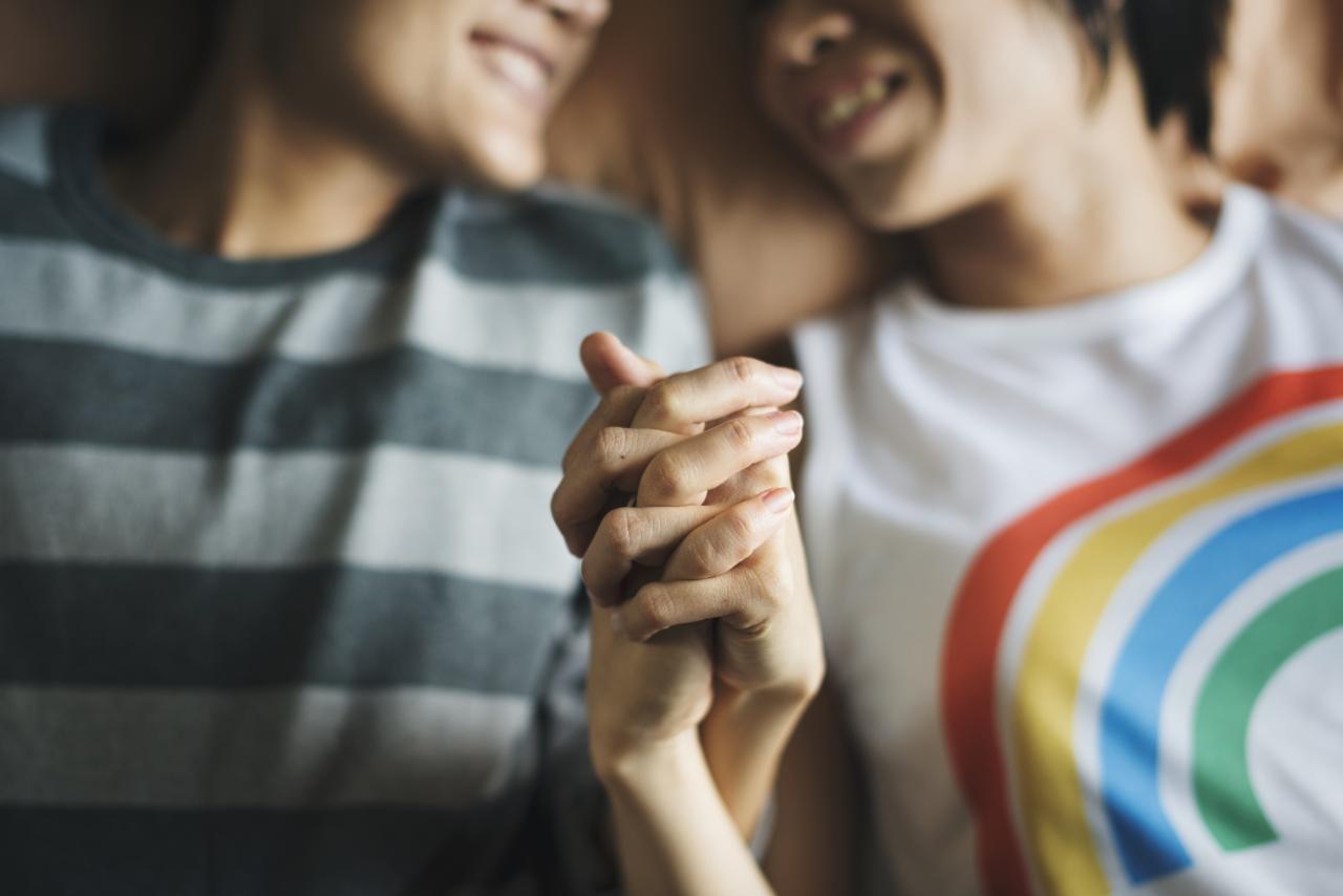 Closeup of two people laying on a bed holding hands, one of them is wearing a rainbow tshirt