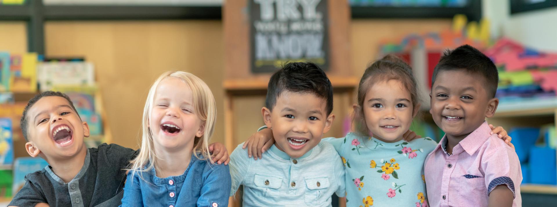 Children sitting in a line with their arms around each other. The kids are laughing and smiling directly at the camera.