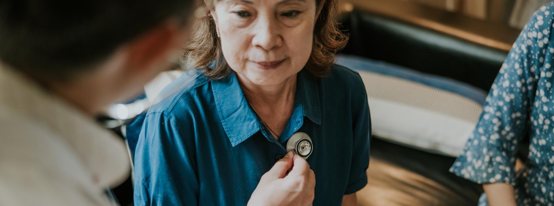 An unknown doctor placing a stethoscope on an older persons chest while their family member sits next to them