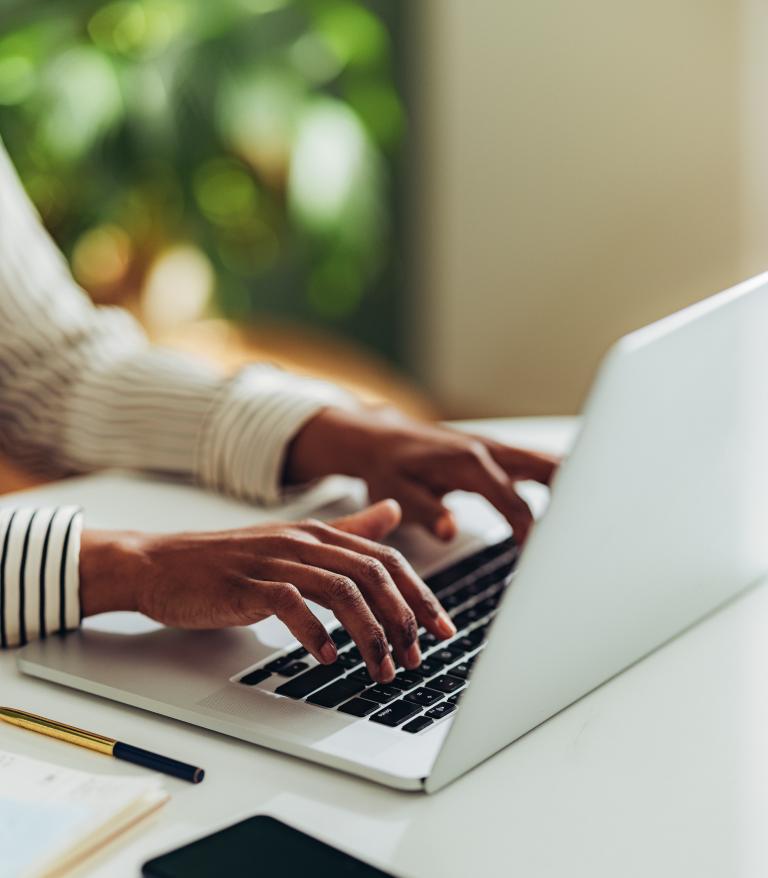 Person typing on a laptop on a white desk with a phone and notepage.