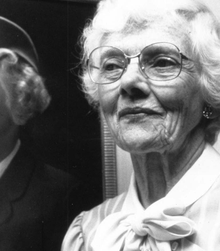 Mary Pack as an older adult standing in front of a photo of herself when she was younger.