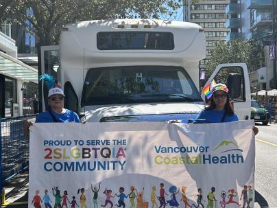 Two VCH employees hold a banner in front of a bus at the 2024 Vancouver pride parade