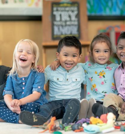 Children sitting in a line with their arms around each other. The kids are laughing and smiling directly at the camera.